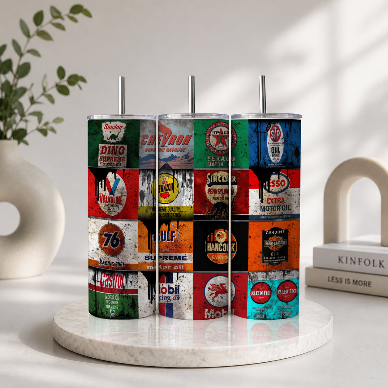 Three metal tumblers with straws feature a colorful collage of vintage gas and oil signs displayed on a marble round stand, promoting the Vintage Gas & Oil Signs Collage Tumbler – Retro Automotive Garage Drinkware.