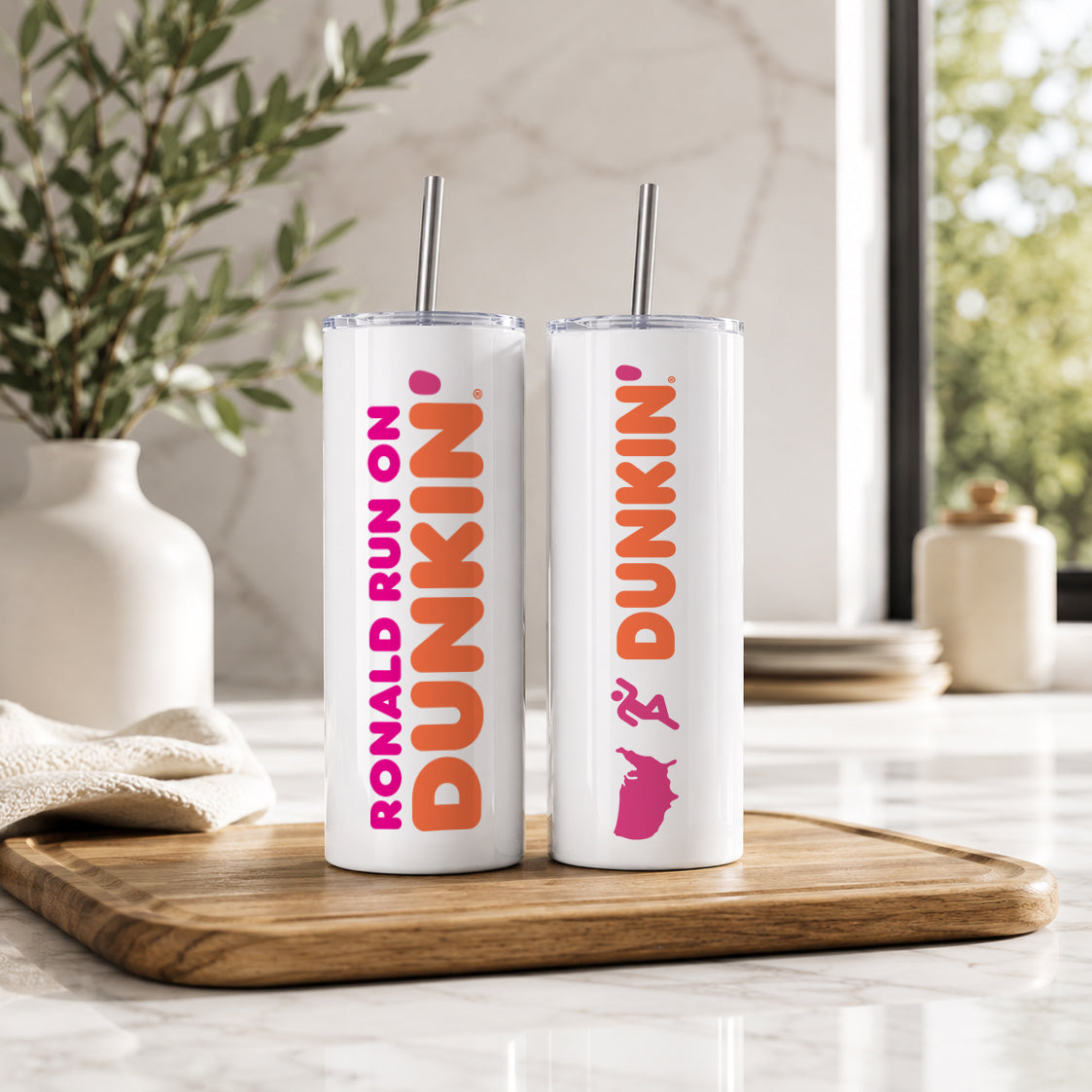 Two white insulated tumblers with metal straws featuring vibrant pink and orange Dunkin style custom designs, displayed on a wooden tray in a modern kitchen setting, showcasing the Personalized Name Tumbler – Custom Dunkin Style Insulated Cup with Straw | Custom Name Gift.