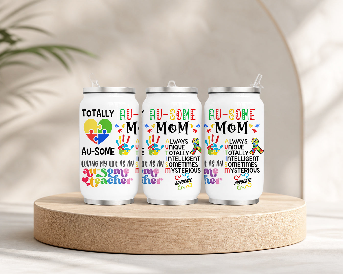 Three white tumblers with colorful puzzle piece designs and text promoting autism awareness, featuring the phrase Proud Au-some Mom Tumbler – Austism Awareness Colorful Puzzle Drinkware, displayed on a wooden platform with a neutral background.